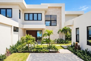 A modern two-story house with a central entrance, a paved driveway, and a well-maintained garden with palm trees. - Beach Front Staffed 6br Villa | Lux Resort by Bocobay (St. Ann’s Bay)