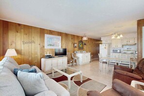 A living room with a sofa, coffee table, and TV, a dining area with chairs, and a kitchen with cabinets and appliances. - The Bounty W1 (Ocean City)