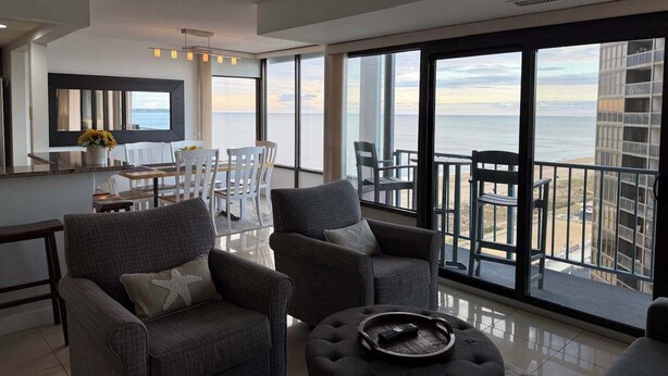 A modern living room with a large balcony, a dining area, and a view of the ocean. - Upscale ocean view condo with pool access & balcony - walk to beach, restaurants (Ocean City)