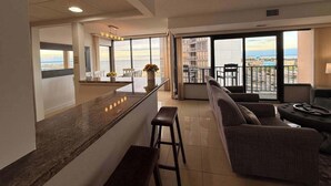 A modern living room with a kitchen area, large windows, and a balcony with a view. - Upscale ocean view condo with pool access & balcony - walk to beach, restaurants (Ocean City)