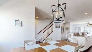 Ein Essbereich mit Holztisch, weißen Stühlen, einer geometrischen Pendelleuchte und einer Küche im Hintergrund.