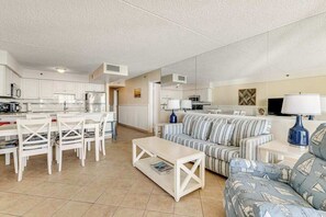 A modern living room with a sofa, coffee table, and dining area with chairs and a kitchen in the background. - Beautiful beachfront condo with pools, ice rink, arcade, gym, & fast WiFi (Ocean City)