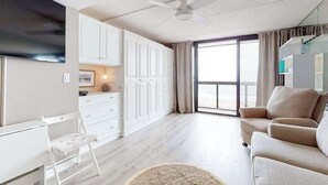 A modern living room with a sofa, a dining area with white cabinets, a TV, and a large window. - Beachfront studio with balcony, AC & beachfront & rooftop pools (Ocean City)