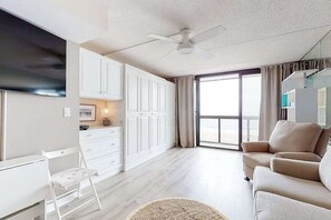 A modern living room with a sofa, a dining area with white cabinets, a TV, and a large window. - Beachfront studio with balcony, AC & beachfront & rooftop pools (Ocean City)