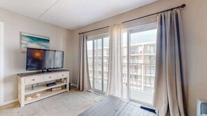 A room with a television on a white cabinet, a large window with curtains, and a view of buildings outside. - Wight Bay South 449 (Ocean City)