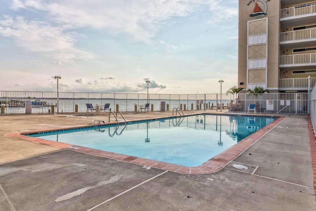 A swimming pool with a view of the ocean, a fenced deck area, and a building with a sailboat logo.