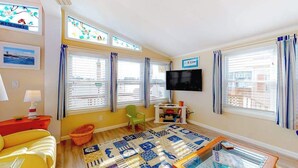 A living room with a yellow sofa, a TV, a bookshelf, and a window with curtains. - Canal front home with free WiFi, deck, private dock, shared pool & tennis (Ocean City)