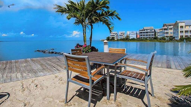 Een houten terras met een tafel en stoelen, palmbomen en uitzicht op de oceaan en gebouwen.