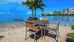 Eine Holzterrasse mit Tisch und Stühlen, Palmen und Blick auf das Meer und die Gebäude.