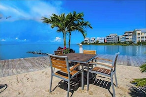 Eine Holzterrasse mit Tisch und Stühlen, Palmen und Blick auf das Meer und die Gebäude.