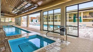 Area piscina interna con pareti in vetro trasparente, soffitto in legno e vista sugli edifici esterni.