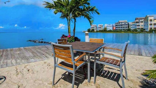A wooden deck with a table and chairs, palm trees, and a view of the ocean and buildings. - Sunset Island - 59 Island Edge (Ocean City)