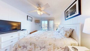 Ein Schlafzimmer mit einem Bett, einem Fernseher, einem Deckenventilator, einem Fenster und einem gerahmten Bild an der Wand.
