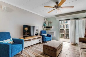 Ein Wohnzimmer mit Flachbildfernseher, zwei blauen Sesseln, einem braunen Hocker und einem Deckenventilator.