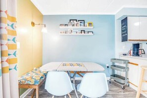 A room with a table, chairs, a bench, a shelf with framed photos, a wall-mounted light, and a kitchen area with cabinets and a coffee machine. - Summer Palace 7 (Ocean City)