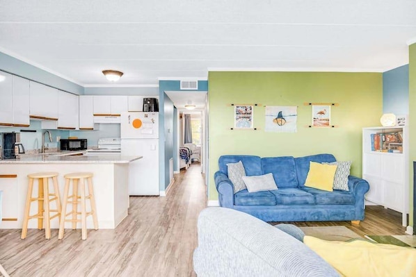 A modern kitchen with white cabinets, a central island with stools, and a blue sofa in a living area with green walls. - Summer Palace 7 (Ocean City)
