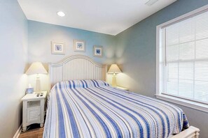 A bedroom with a bed, two bedside tables with lamps, a window with blinds, and three framed pictures on the wall. - Captain's View 2 (Ocean City)