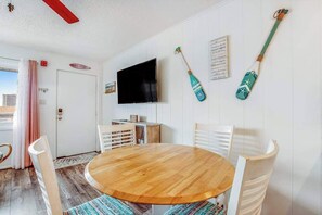 Ein Zimmer mit einem runden Holztisch, weißen Stühlen, einem an der Wand montierten Fernseher und einem Surfbrett, das an der Wand hängt.