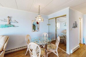 A dining area with a round glass table, white wicker chairs, a chandelier, and wall decorations including a lighthouse and a mirror. - Oceanfront Condo with Balcony, Sunrise Views, Free WiFi & Washer/Dryer (Ocean City)