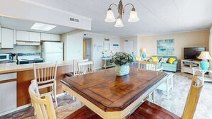 A modern kitchen with white cabinets, a dining area with a wooden table, and a living room with a sofa and TV. - Ocean Place 519 (Ocean City)