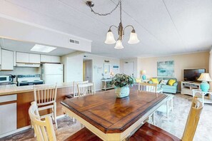 Eine moderne Küche mit weißen Schränken, ein Essbereich mit Holztisch sowie ein Wohnzimmer mit Sofa und Fernseher.