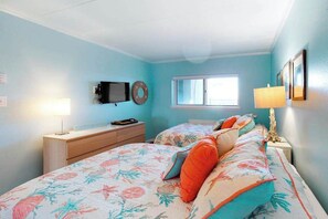Ein Schlafzimmer mit zwei Betten, einem Fernseher, einem Fenster und einem Schreibtisch mit einer Lampe.