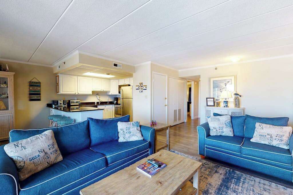 A living room with two blue sofas, a wooden coffee table, and a kitchen in the background.