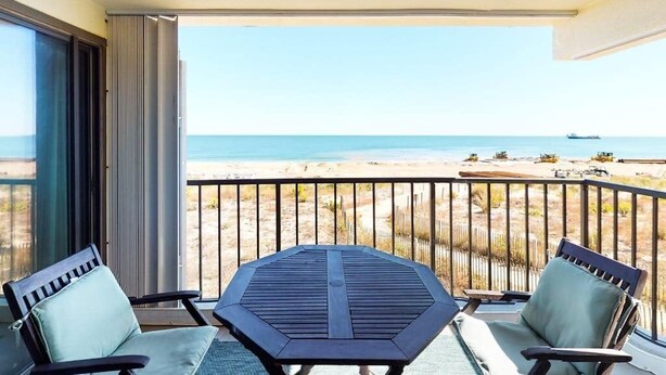 A balcony with a table and chairs, overlooking a beach with a fence and a ship in the distance. - Waikiki 102 (Ocean City)