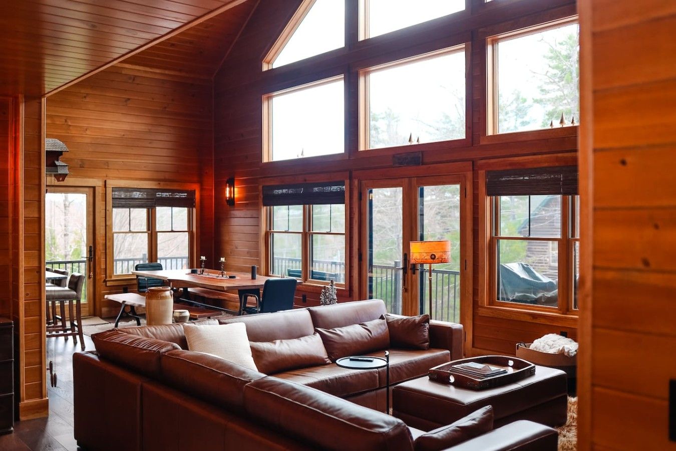 A cozy cabin interior with a large wooden ceiling, a leather sofa, a dining table, and multiple windows.