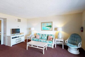 A hotel room with a white entertainment unit, a floral sofa, a round chair, and a painting of a beach scene. - Simple, Oceanfront Condo with Central AC, Free WiFi & Easy Beach Access (Ocean City)