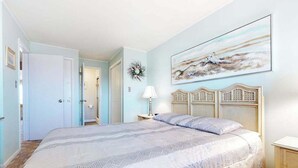 A bedroom with a bed, a nightstand, a lamp, a wall-mounted painting, and a door. - Thunder Island 28A (Ocean City)