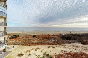 Ein Strandgrundstück mit Blick auf das Meer, ein sandiger Bereich mit spärlicher Vegetation und ein mehrstöckiges Gebäude mit Balkonen.