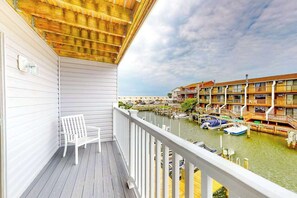 Un balcón con una silla blanca, techo de madera y vistas a un puerto deportivo con barcos y edificios.