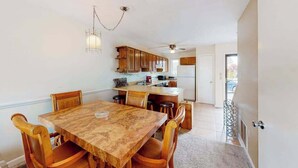 A dining area with a wooden table and chairs, a kitchen in the background, and a ceiling fan. - Dawn's Cove F (Ocean City)