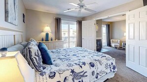 A bedroom with a bed, a ceiling fan, a desk with a lamp, a chair, and a door leading to another room. - Lucayan 18 (Ocean City)