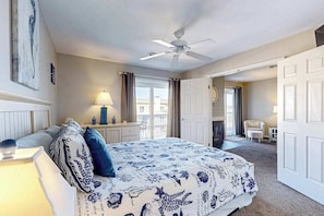 A bedroom with a bed, a ceiling fan, a desk with a lamp, a chair, and a door leading to another room. - Lucayan 18 (Ocean City)