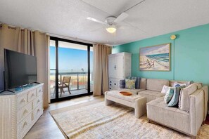 A living room with a TV, sofa, and a painting of a beach scene. - Condo in Ocean City with pools, gym, & game room (Ocean City)