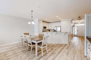 A modern kitchen with white cabinets, a central island, and a dining area with a wooden table and chairs. - Stylish, single-level condo with pools, a picnic area, & private beach access (Ocean City)