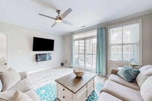 A living room with a ceiling fan, a flat-screen TV mounted on the wall, a wooden coffee table, and a sofa with a patterned rug. - Stylish, single-level condo with pools, a picnic area, & private beach access (Ocean City)