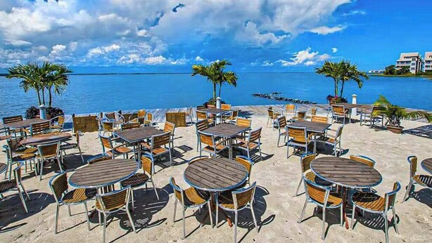 A beachside café with round tables and chairs, palm trees, and a view of the sea. - Coastal retreat with beach and shared pool - balconies, AC, & W/D (Ocean City)