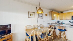 A dining area with a wooden table and chairs, a television, and a kitchen with wooden cabinets. - Oceanfront escape close to boardwalk with balcony, W/D, and direct beach access (Ocean City)