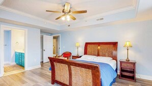 Quarto com cama de madeira, duas luminárias de cabeceira, ventoinha no teto e porta para outro cômodo.