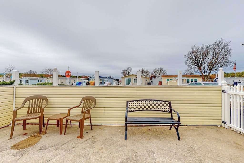 A fenced outdoor area with a bench and two chairs, a stop sign, and a row of houses in the background.