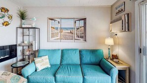 Ein blaugrünes Sofa, ein Fernseher, ein Fenster mit Blick auf den Strand, eine Lampe und ein Regal mit dekorativen Gegenständen.