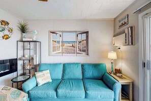 A teal sofa, a TV, a window with a beach view, a lamp, and a shelf with decorative items. - Renovated bayfront condo with water-view balcony & fast WiFi - walk to beach (Ocean City)
