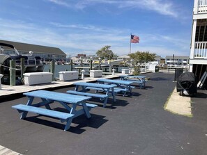 A marina with blue picnic tables, a docked boat, and an American flag. - Renovated bayfront condo with water-view balcony & fast WiFi - walk to beach (Ocean City)