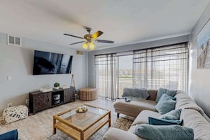 A living room with a sofa, a coffee table, a TV, and a ceiling fan. - Bayside condo with balcony, sunset view, pool & tennis - steps to Jolly Roger (Ocean City)