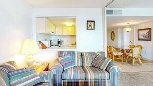A living room with a sofa, a coffee table, a lamp, a dining area with chairs, and a kitchen visible through an open door. - Fountainhead Towers 305 (Ocean City)