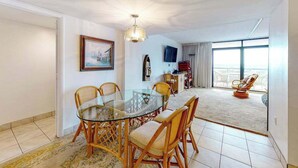 A dining area with a glass table and wicker chairs, a painting on the wall, a television, and a view through large windows. - Fountainhead Towers 305 (Ocean City)