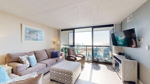 A modern living room with a sofa, ottoman, and chairs, a flat-screen TV, and a balcony with a view. - Family-friendly oceanfront condo with balcony, pools, game room & gym (Ocean City)